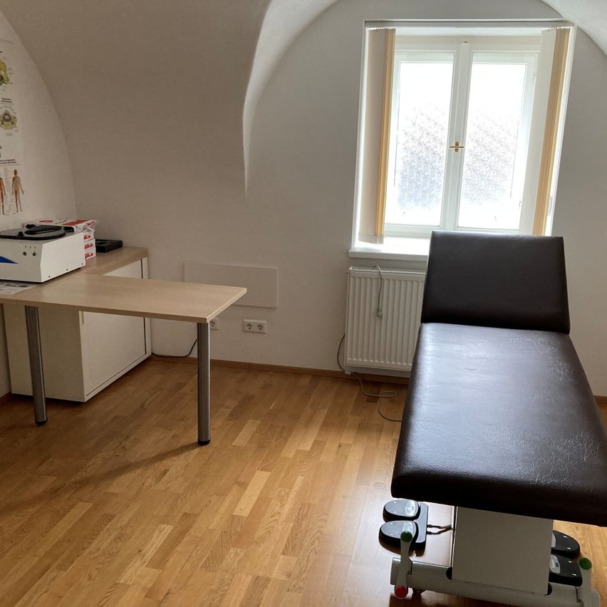 Ein kleiner Untersuchungsraum mit Holzboden, einem Schreibtisch mit einer Maschine, einem Fenster mit weißem Rahmen und einem schwarzen Lederuntersuchungstisch.
