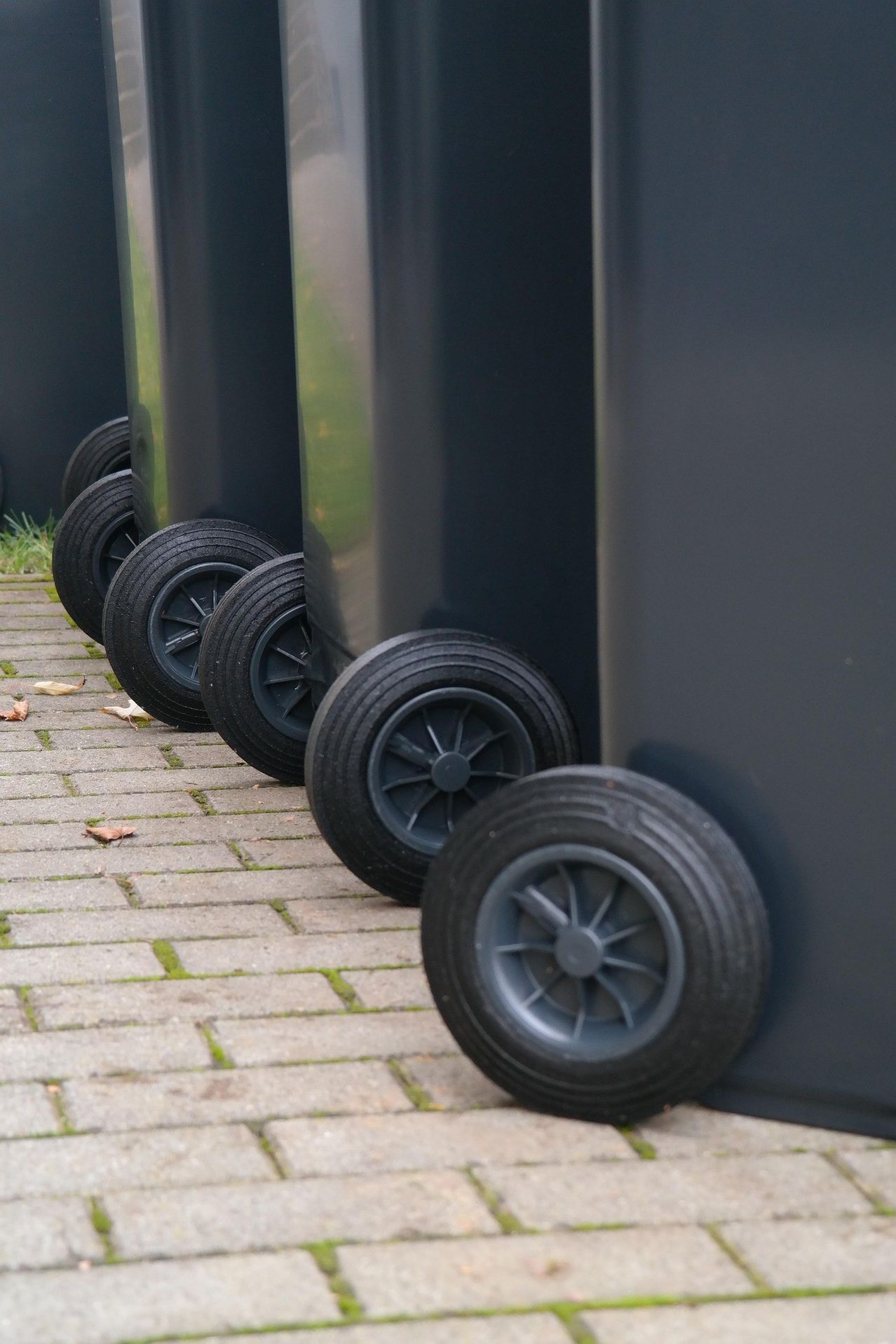 Vier große schwarze Behälter mit Rädern sind auf einem Ziegelweg aufgereiht. Die Behälter sind zylindrisch und die Räder sind schwarz. Hinter den Behältern befindet sich eine Grasfläche.