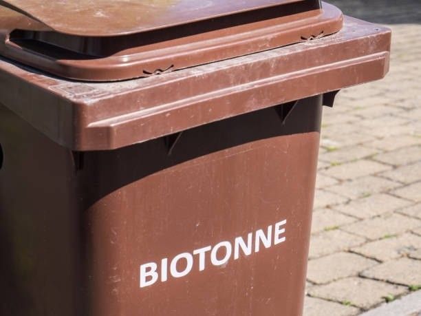 Ein brauner Mülleimer mit offenem Deckel, beschriftet mit 'BIOTONNE', steht auf einem gepflasterten Weg.