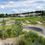 Pumptrack St. Ruprecht-Logo. Eine Luftaufnahme eines Skateparks mit Bäumen, Blumen und Häusern in der Ferne. Der Park hat Rampen und Kurven auf einer Schotteroberfläche.