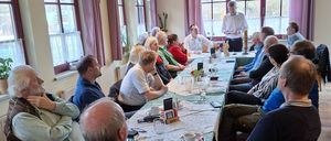 Eine Gruppe von Menschen sitzt um einen Tisch in einem Esszimmer, möglicherweise in einer Besprechung. Sie blicken auf die Person, die vor ihnen steht und spricht. Der Raum hat Glasfenster mit Vorhängen.