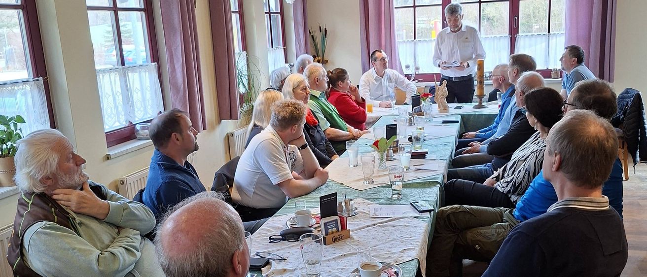 Eine Gruppe von Menschen sitzt um einen Tisch in einem Esszimmer, möglicherweise in einer Besprechung. Sie blicken auf die Person, die vor ihnen steht und spricht. Der Raum hat Glasfenster mit Vorhängen.
