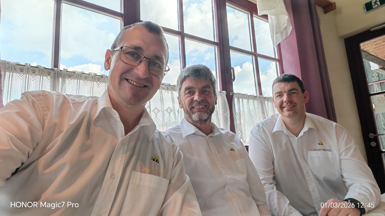 Drei Männer lächeln und posieren für ein Foto vor einem Fenster mit Vorhängen. Der Mann auf der linken Seite trägt eine Brille. Der mittlere Mann hat einen Bart und ein Logo auf seinem Hemd. Der Mann auf der rechten Seite hat kurze Haare.