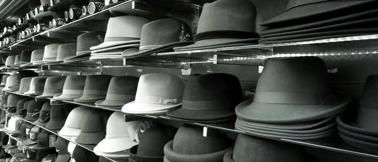 Ein schwarz-weißes Bild zeigt die Hutauswahl eines Ladens mit Hüten in verschiedenen Stilen und Farben, von Fedoras bis hin zu Baseballkappen, ordentlich auf Regalen angeordnet.