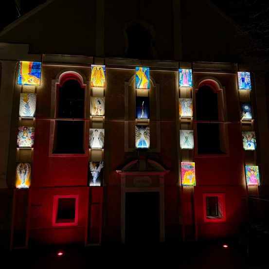 Ein Gebäude mit mehreren beleuchteten Tafeln, auf denen jeweils eine einzigartige Engelsfigur abgebildet ist, wird bei Nacht durch rote Beleuchtung hervorgehoben.