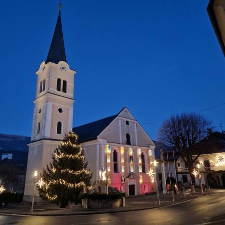 Eine Kirche mit Kirchturm und Uhr steht hell erleuchtet in der Dämmerung. Ein mit Lichtern geschmückter Weihnachtsbaum steht davor. Das Gebäude hat bunte Fenster und pinke Beleuchtung.