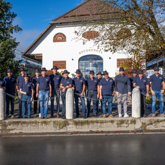 Eine Gruppe von Männern in identischen Uniformen steht vor einem Gebäude mit dem Schild 'ÖTZSTAL'. Sie sind mit Metallketten aneinander gefesselt.