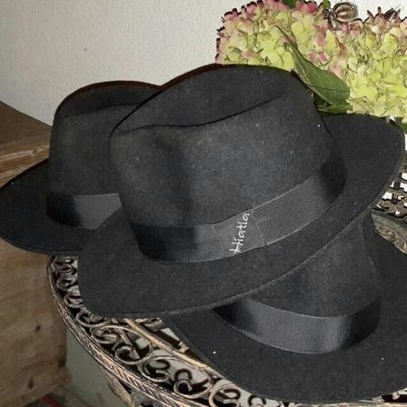 Zwei schwarze Hüte mit schwarzen Bändern liegen auf einem Tisch, im Hintergrund befindet sich ein Blumenstrauß.