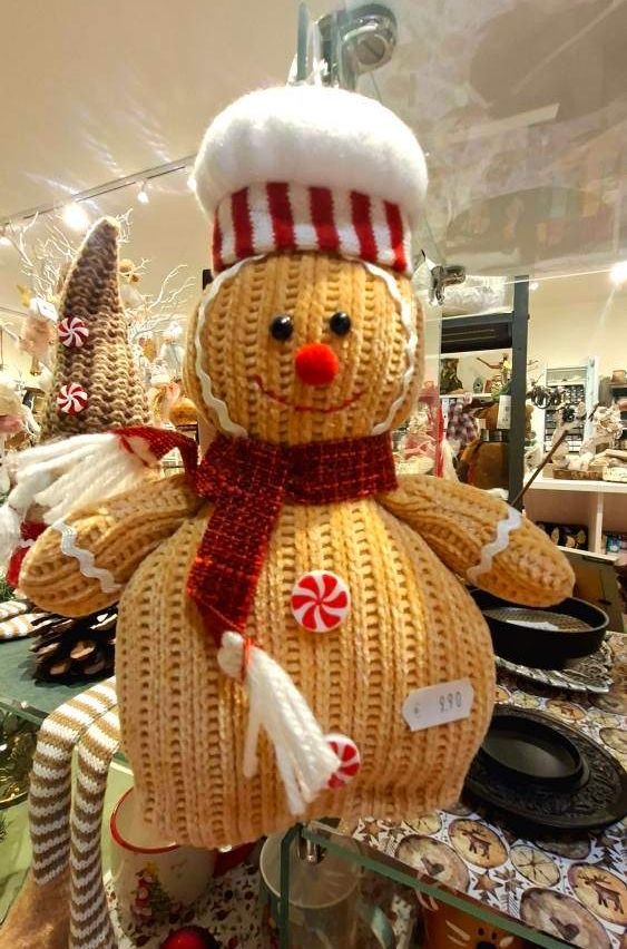 Ein gestricktes Lebkuchenmännchen mit einem rot-weiß gestreiften Hut, einem roten Schal und einer weißen Zuckerstange wird in einem Geschäft ausgestellt.