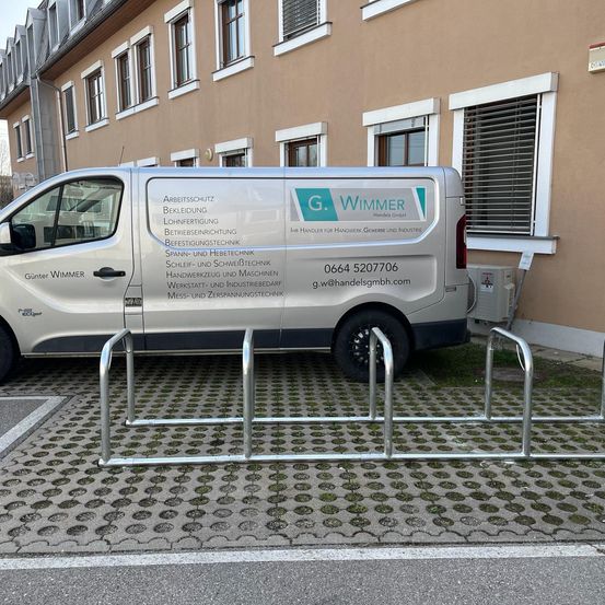 Ein silberner Van mit G. Wimmer Schriftzug steht neben einem Gebäude mit mehreren Fenstern und einem Metallfahrradständer davor.
