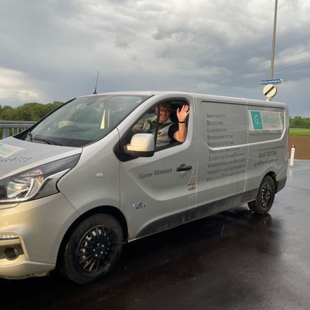 Ein silberner Lieferwagen mit 'G. Wimmer' auf der Seite, gefahren von einer winkenden Person, parkt auf einer nassen Straße unter einem bewölkten Himmel.