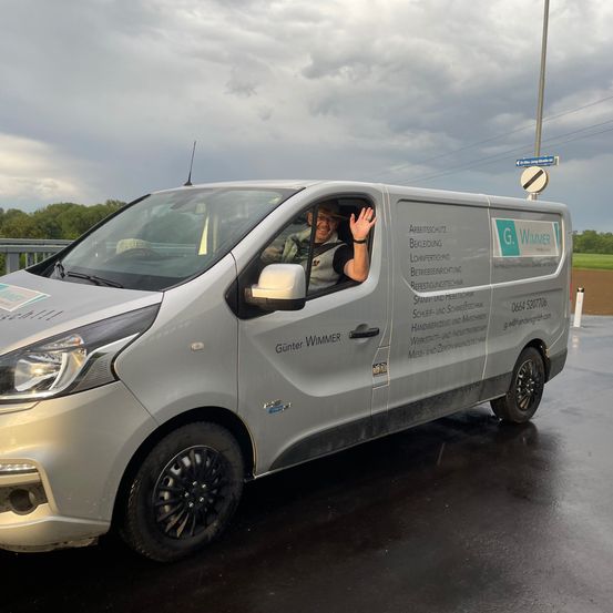 Ein silberner Lieferwagen mit 'G. Wimmer' auf der Seite, gefahren von einer winkenden Person, parkt auf einer nassen Straße unter einem bewölkten Himmel.