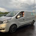 Ein silberner Lieferwagen mit dem Namen Gunter Wimmer an der Seite ist auf einer nassen Straße geparkt. Ein Mann im Inneren des Lieferwagens winkt. Der Himmel ist bewölkt.