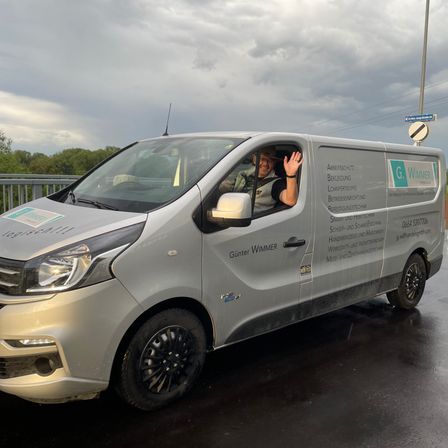 Ein silberner Lieferwagen mit dem Namen Gunter Wimmer an der Seite ist auf einer nassen Straße geparkt. Ein Mann im Inneren des Lieferwagens winkt. Der Himmel ist bewölkt.