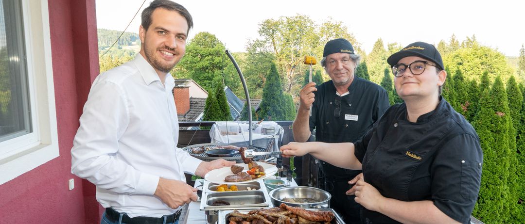 Drei Personen stehen hinter einem Tisch voller verschiedenes Fleisch und Gemüse, wahrscheinlich in Vorbereitung auf ein Grillevent. Der Mann auf der linken Seite lächelt und hält einen Teller, während die beiden anderen Lebensmittel grillen.