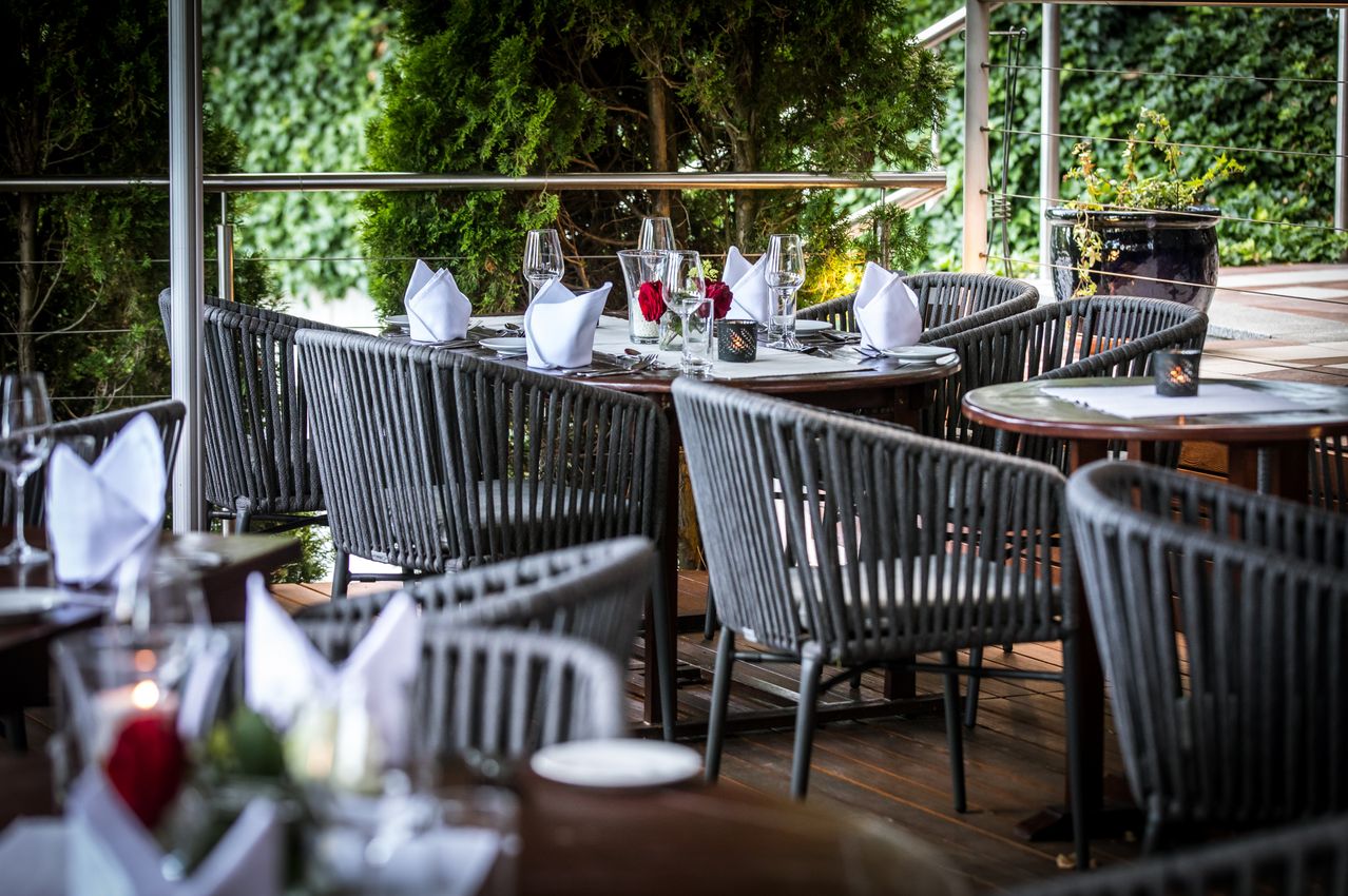 Ein Outdoor-Restauranttisch, der für eine Mahlzeit mit Weingläsern, Servietten und einer Tischdekoration aus roten Rosen vorbereitet ist. Der Tisch ist von Korbstühlen umgeben.