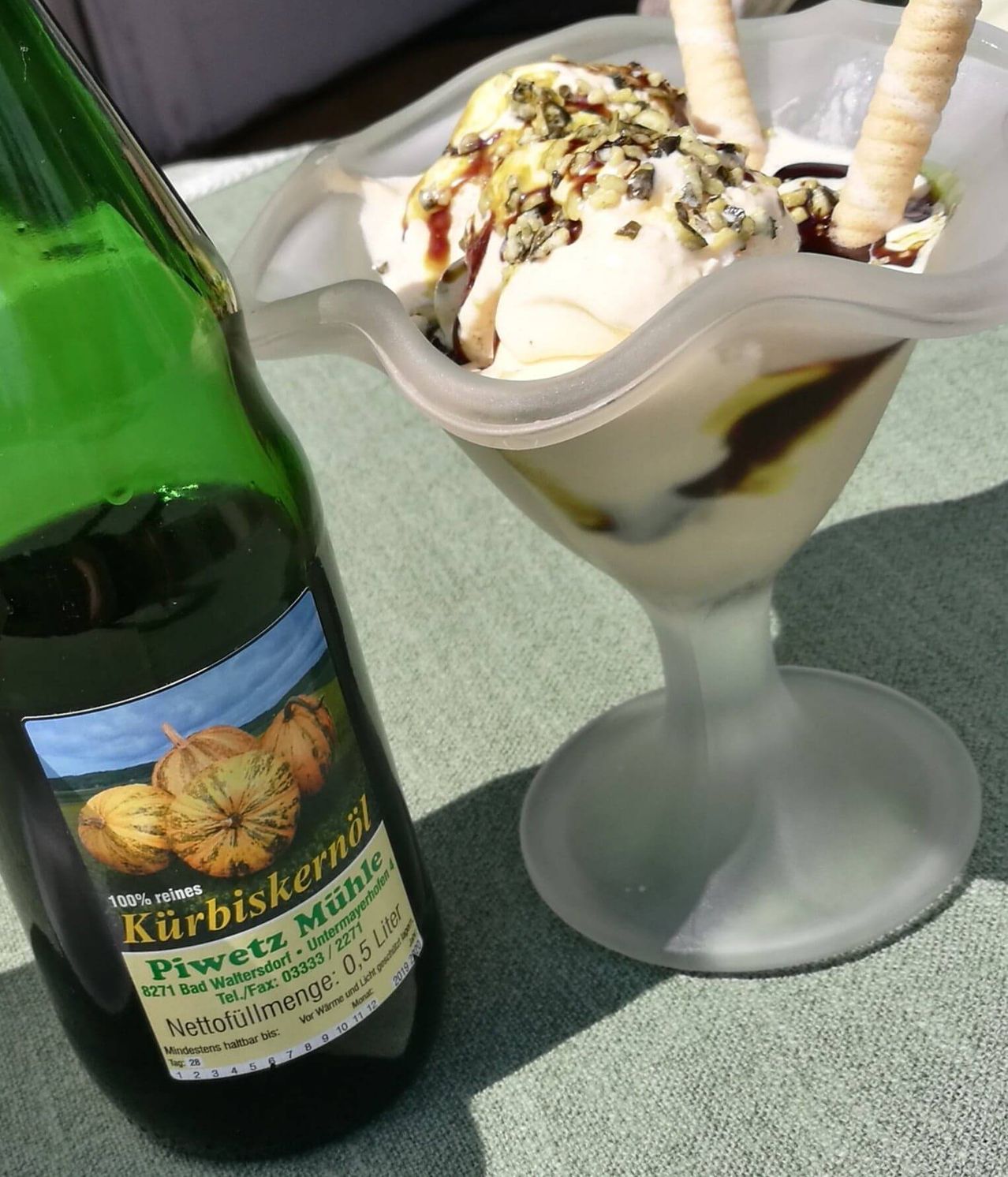Eine Flasche Kürbiskernöl neben einem Glas Eis mit Schokolade und Nüssen.
