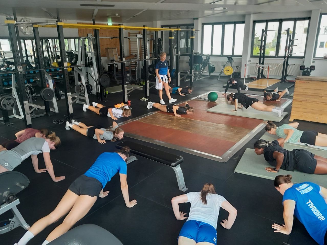 Eine Gruppe von Menschen in einem Fitnessstudio, die Bodenübungen mit einem Trainer durchführt. Im Hintergrund sind verschiedene Fitnessgeräte zu sehen.