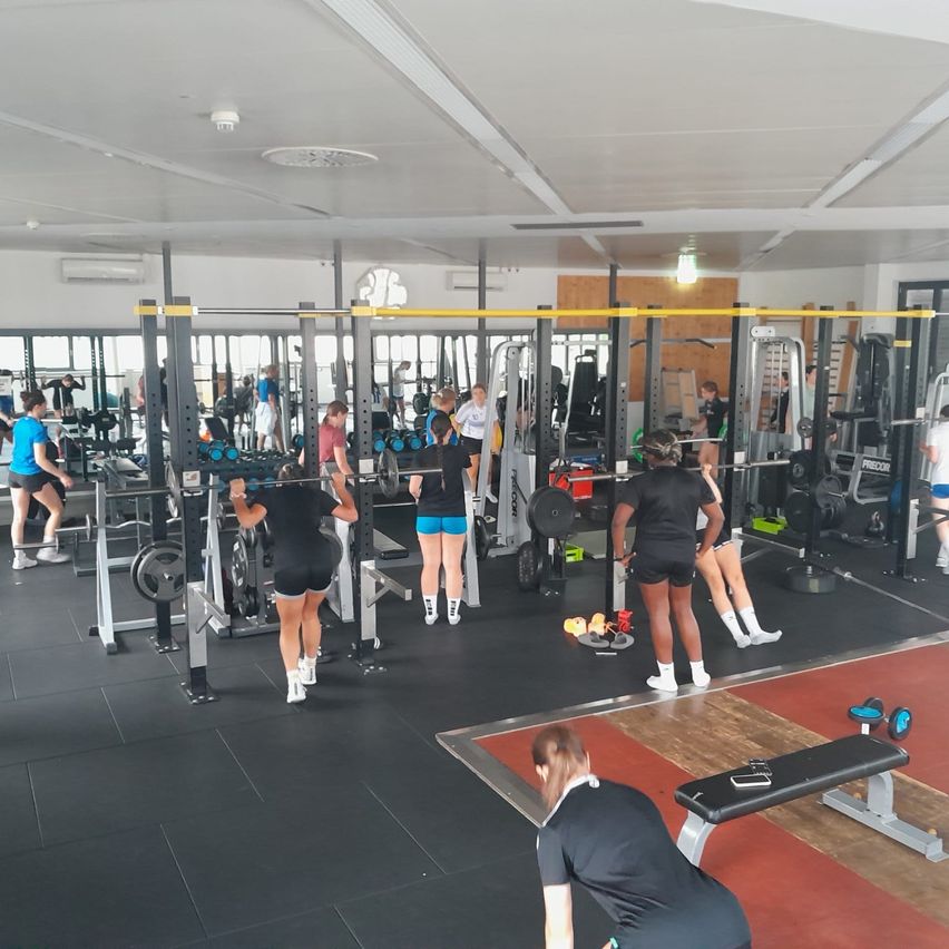 Ein Fitnessstudio mit mehreren Personen, die verschiedene Geräte benutzen. Einige machen Kniebeugen, während andere Gewichtsmaschinen nutzen.