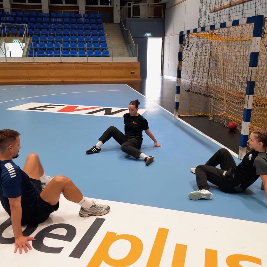 Drei Personen sitzen auf dem Boden einer Sporthalle. Zwei von ihnen tragen schwarze Sportbekleidung und eine trägt Blau. Sie alle schauen auf eine Frau, die in der Mitte steht. Hinter ihnen steht ein Tor mit einem gelben Netz. Der Boden hat ein blaues und weißes Design.