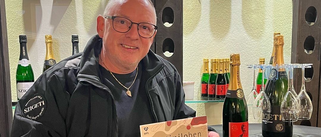 Ein Mann mit Brille und einer schwarzen Jacke steht hinter einem Tisch und hält eine Karte mit dem Aufdruck 'martinilobe' und dem Datum '7.9.november'. Hinter ihm sind Regale mit Weinflaschen zu sehen.