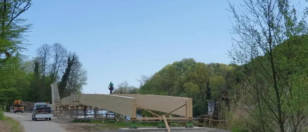 Eine große Holzbrücke wird an einem sonnigen Tag gebaut. Arbeiter sind auf der Brücke, wobei eine Person auf einer Leiter steht. Hinter der Brücke befinden sich Bäume und ein entfernter Berg.