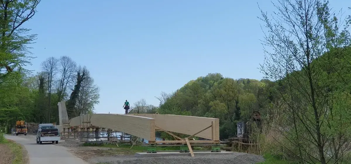 Eine große Holzbrücke wird an einem sonnigen Tag gebaut. Arbeiter sind auf der Brücke, wobei eine Person auf einer Leiter steht. Hinter der Brücke befinden sich Bäume und ein entfernter Berg.