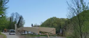 Eine große Holzbrücke wird an einem sonnigen Tag gebaut. Arbeiter sind auf der Brücke, wobei eine Person auf einer Leiter steht. Hinter der Brücke befinden sich Bäume und ein entfernter Berg.