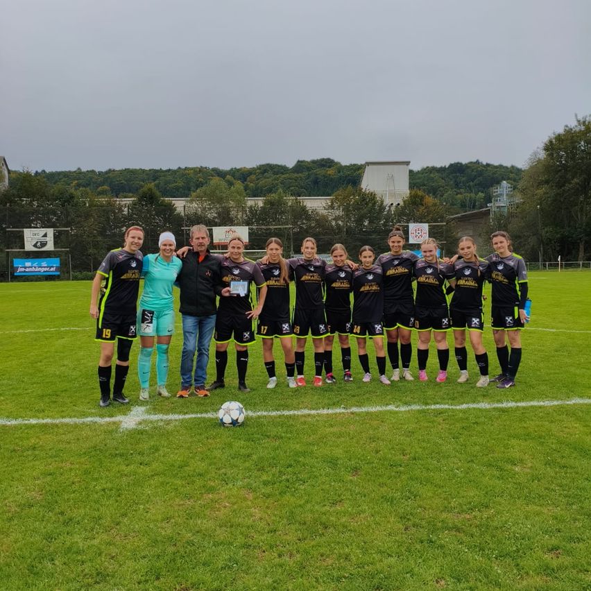 Eine Frauenfußballmannschaft steht auf einem Rasenfeld, zwei Personen halten eine Trophäe. Dahinter befinden sich verschiedene Schilder und Bäume.