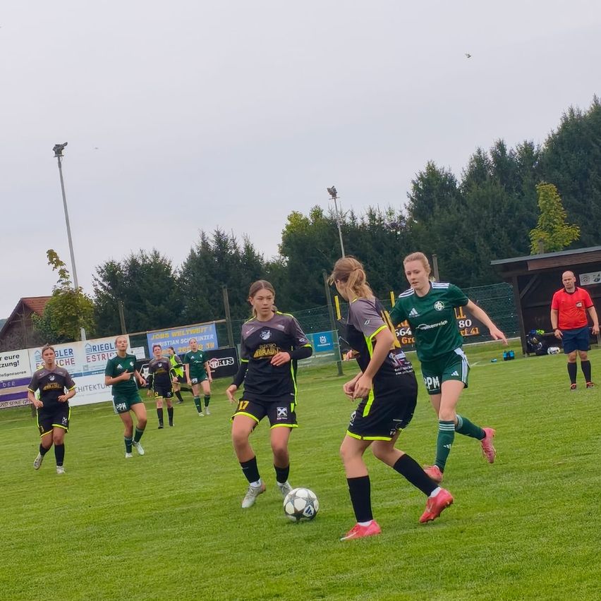Eine Gruppe von Fußballspielerinnen spielt auf einem Feld. Zwei Spielerinnen rennen in Richtung Ball, während andere hinter ihnen herlaufen. Ein Schiedsrichter steht in der Nähe des Torpfostens. Bäume und Werbetafeln befinden sich rund um das Feld.