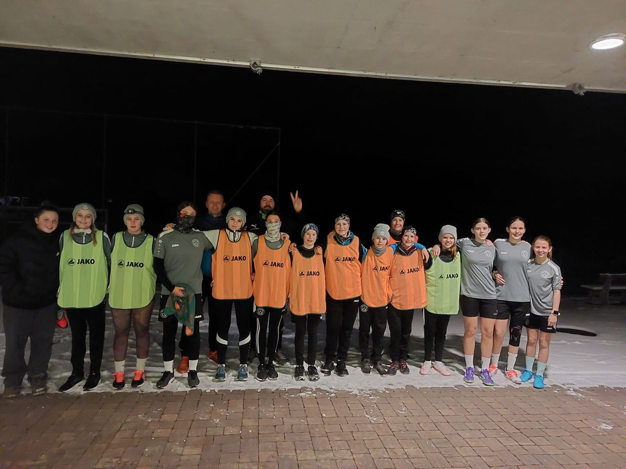 Eine Gruppe von Menschen, gekleidet in Sportausrüstung, posiert für ein Foto auf einer verschneiten Oberfläche. Sie tragen orangefarbene Westen mit dem Wort Jako, einige mit Helmen und Masken. Hinter ihnen befindet sich ein dunkler Bereich und eine große Struktur.