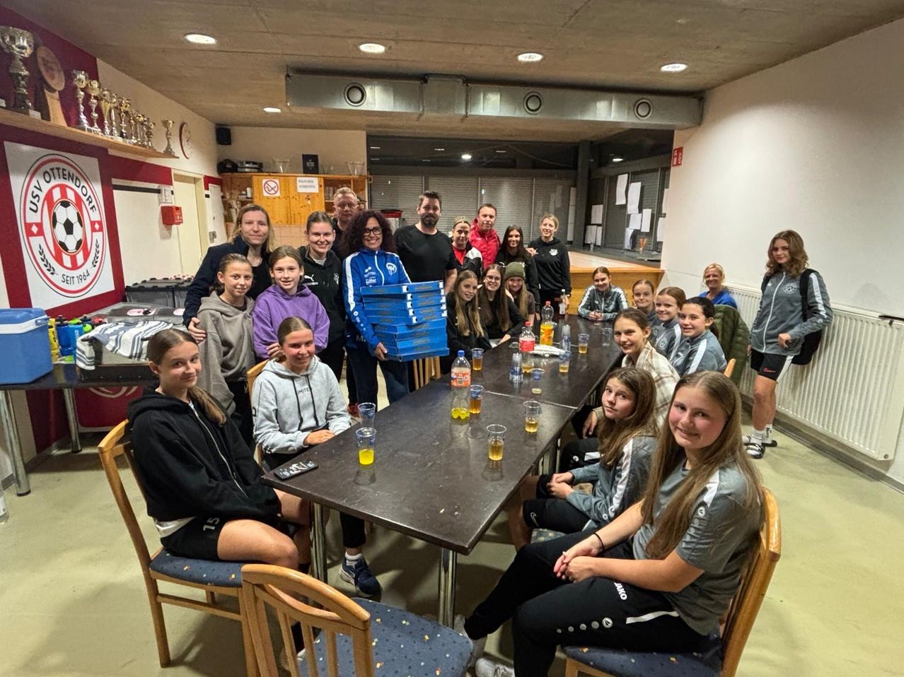 Eine Gruppe junger Athletinnen und Trainer posiert für ein Foto um einen Tisch mit Getränken, in einer Sporteinrichtung.