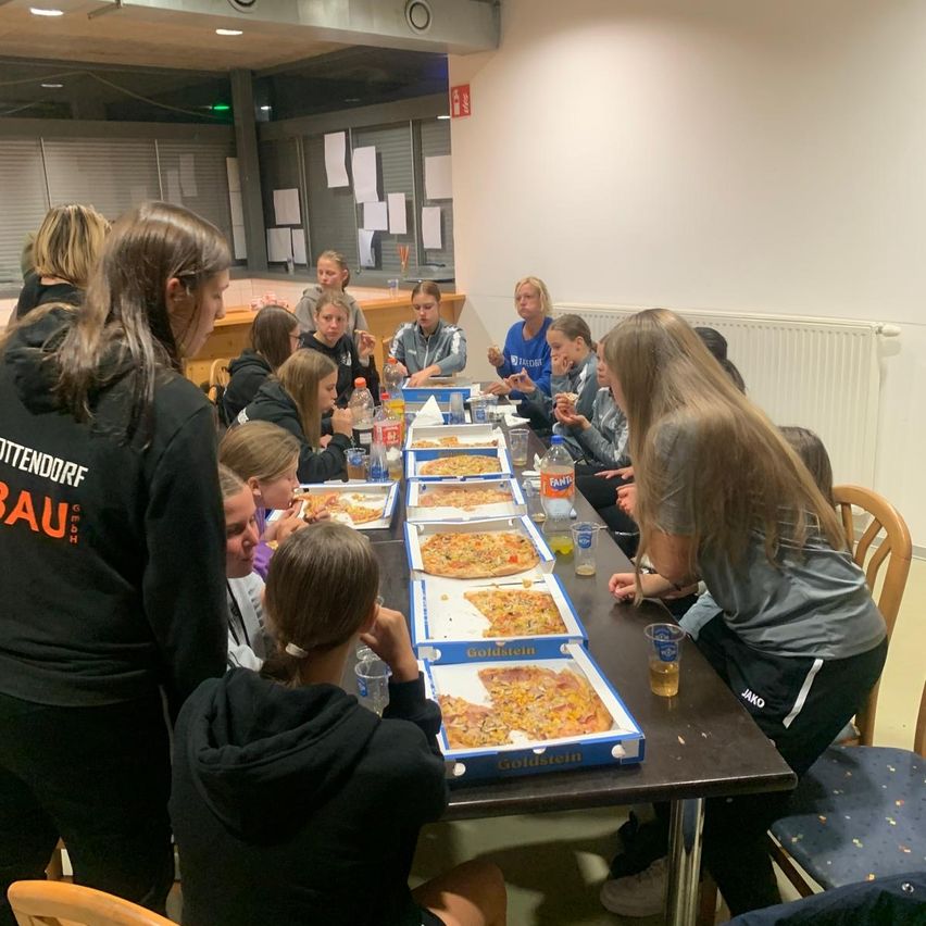 Eine Gruppe von Frauen in Sporttrikots sitzt um einen Tisch in einem Raum und isst Pizza. Sie schauen auf die Pizzakartons und lächeln.