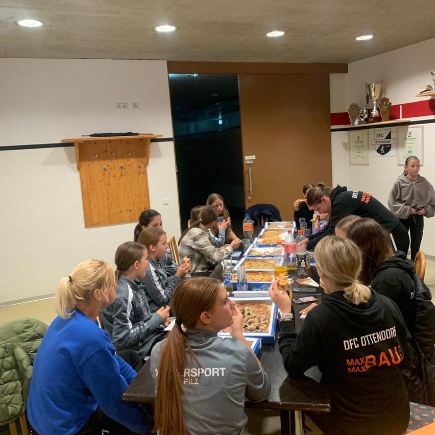 Eine Gruppe junger Frauen in Sweatshirts sitzt um einen Tisch herum und isst Pizza. Eine Person lehnt sich mit einem Pizzastück in der Hand nach vorne. Im Hintergrund eine Tür und ein Trophäenregal.