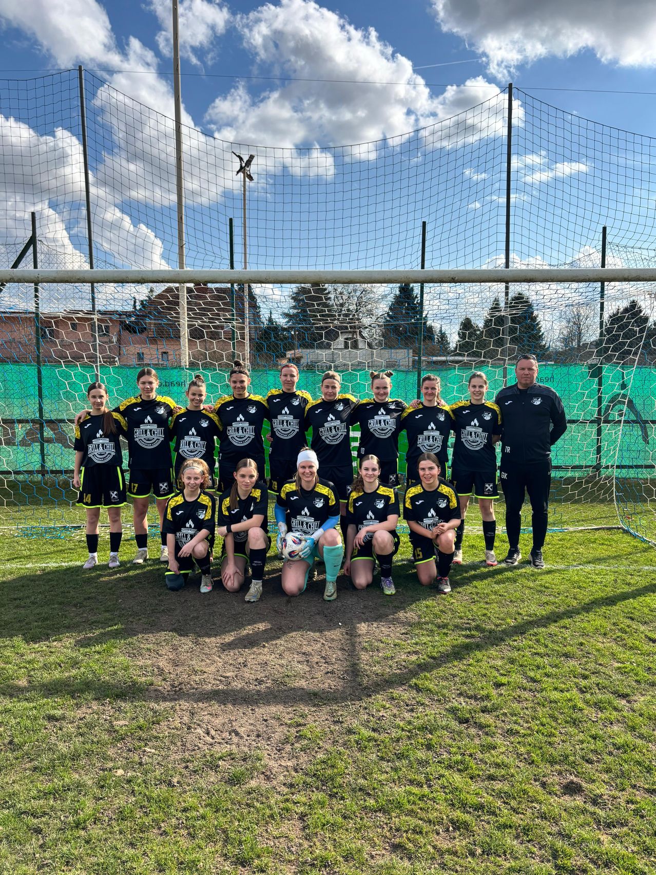 Eine Frauenfußballmannschaft posiert für ein Foto vor einem Fußballtor, mit einem bewölkten Himmel im Hintergrund.