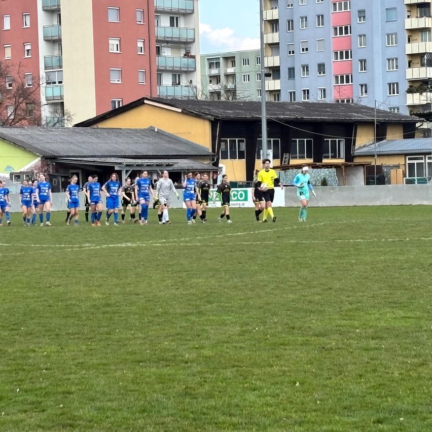 Eine Fußballmannschaft in blauen Uniformen läuft über das Feld, mit einem Schiedsrichter und einem Spieler in Gelb in der Nähe der weißen Linie. Im Hintergrund befinden sich einige Gebäude.