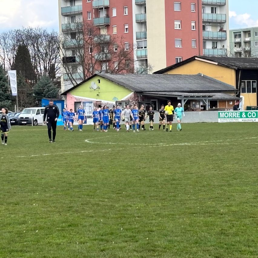 Zwei Fußballmannschaften laufen von einem Rasenfeld, eine in blauen Uniformen und die andere in Schwarz. Das Feld ist von Gebäuden umgeben, von denen eines ein Schild mit der Aufschrift MORRE & CO trägt.