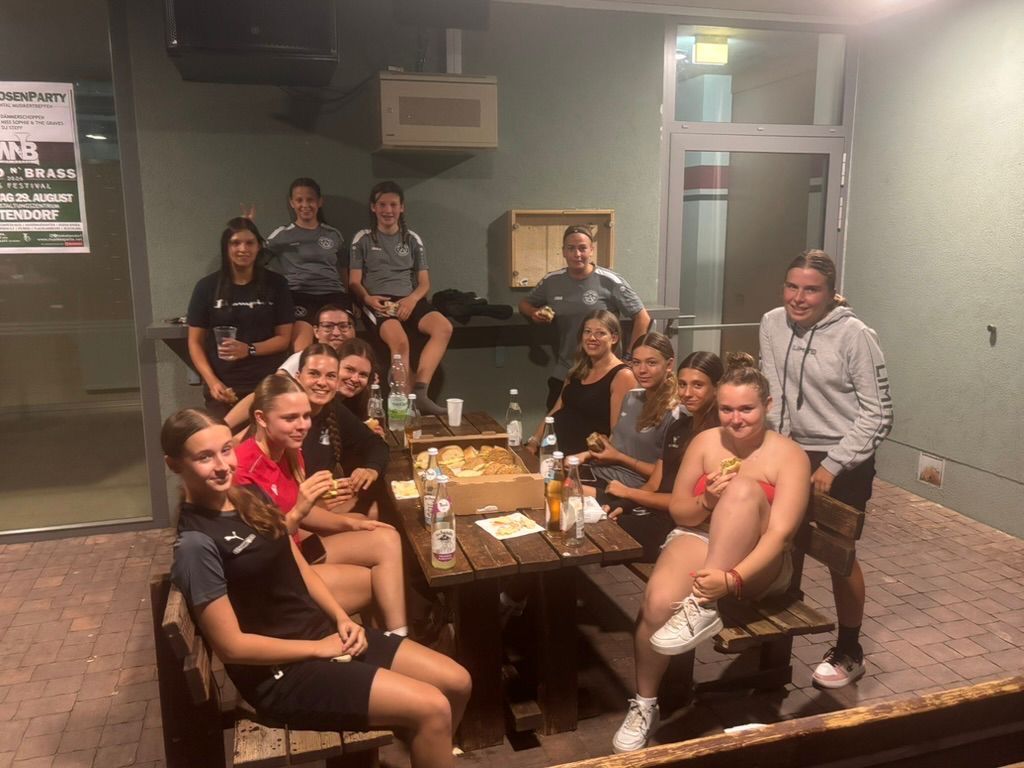 A group of young women in sports attire are gathered around a table, smiling and posing for a photo. The table is laden with food and drinks, and some are eating. They are in a room with a brick wall and a door.