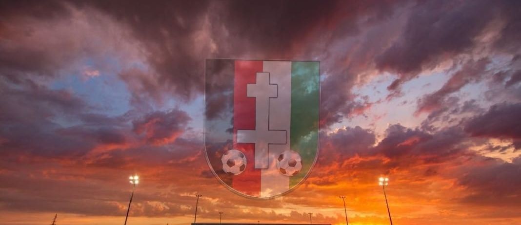 Ein Fußballfeld mit Spielern, unter einem Sonnenuntergangshimmel mit Wolken, hat ein Logo mit zwei Fußballbällen und einem Kreuz.