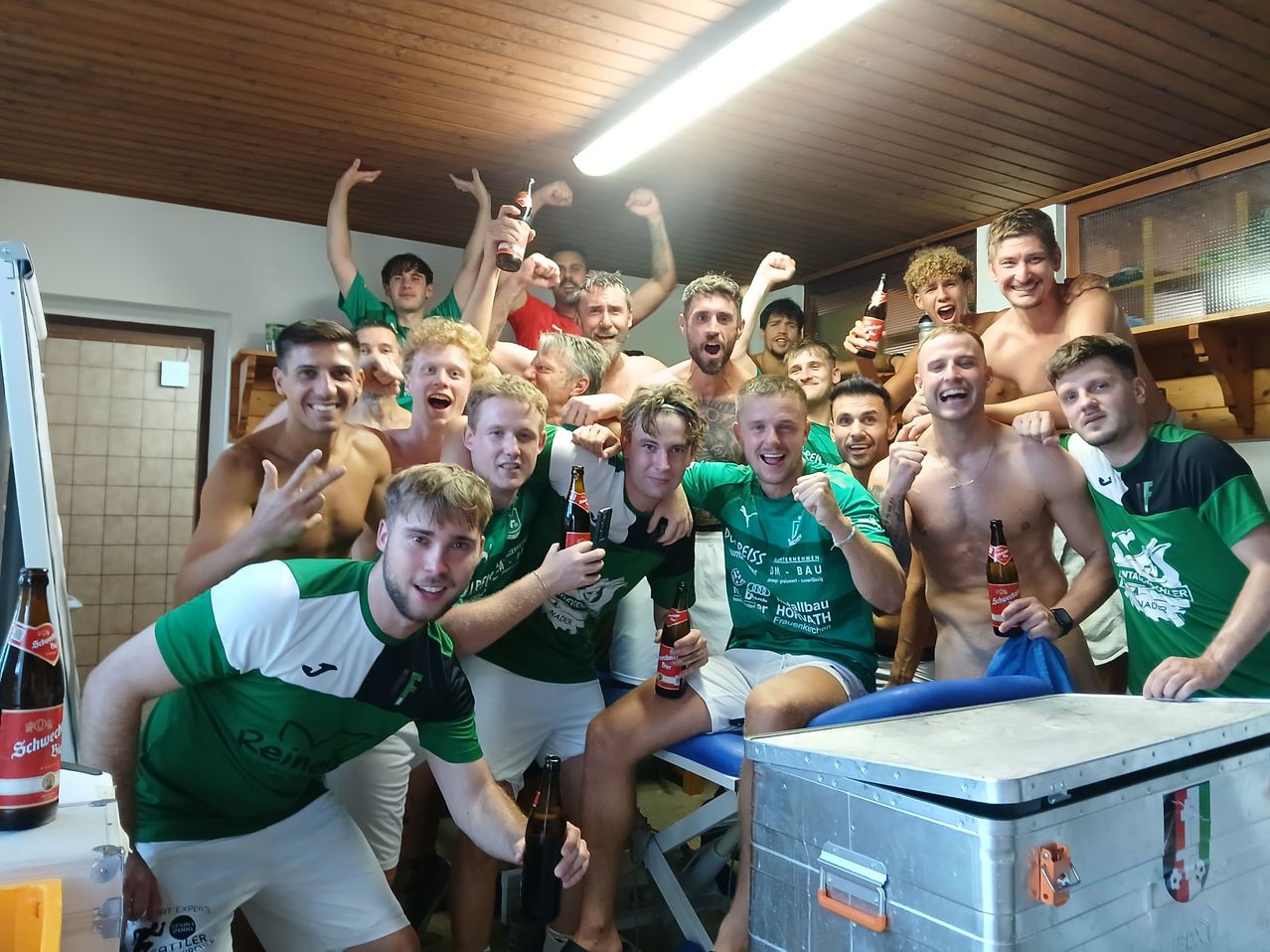 Eine Gruppe junger Männer in grünen und weißen Trikots, teilweise ohne Oberteile, feiert in einer Umkleidekabine mit Bierflaschen, lächelnden Gesichtern und erhobenen Armen.
