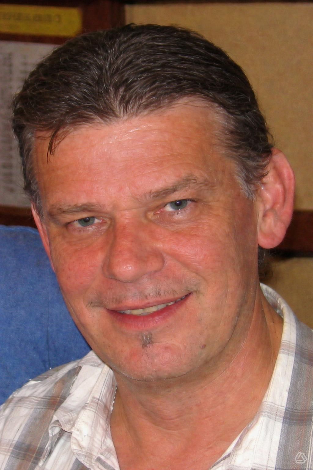 Ein lächelnder Mann mittleren Alters mit kurzen Haaren und einem Schnurrbart steht vor einer gelben Wand und trägt ein weißes Hemd mit grauen Karos.
