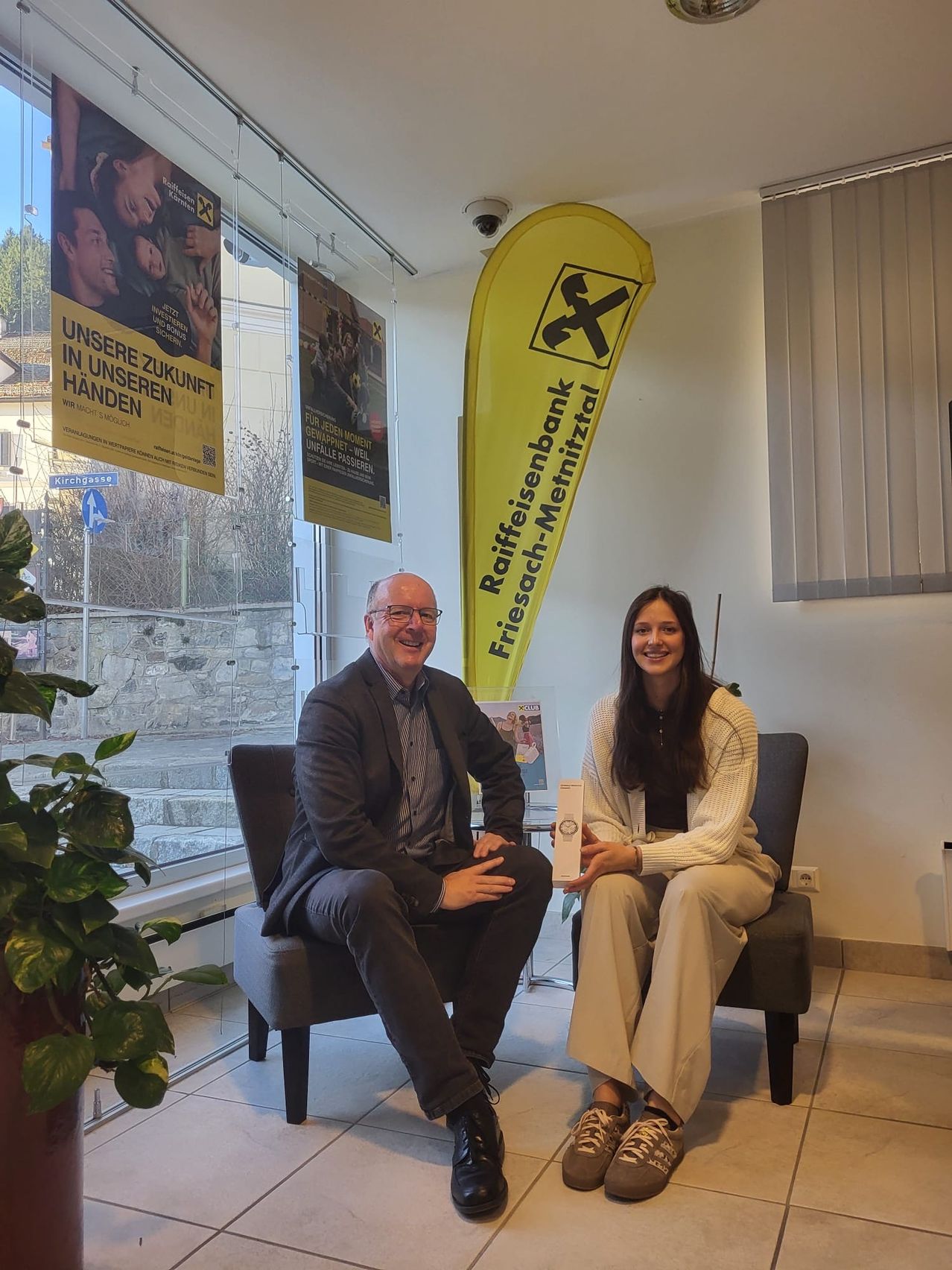 Ein Mann und eine Frau sitzen lächelnd auf Stühlen in einer Lobby. Hinter ihnen befinden sich Plakate und ein Banner. Links stehen eine Pflanze und ein Glasfenster.