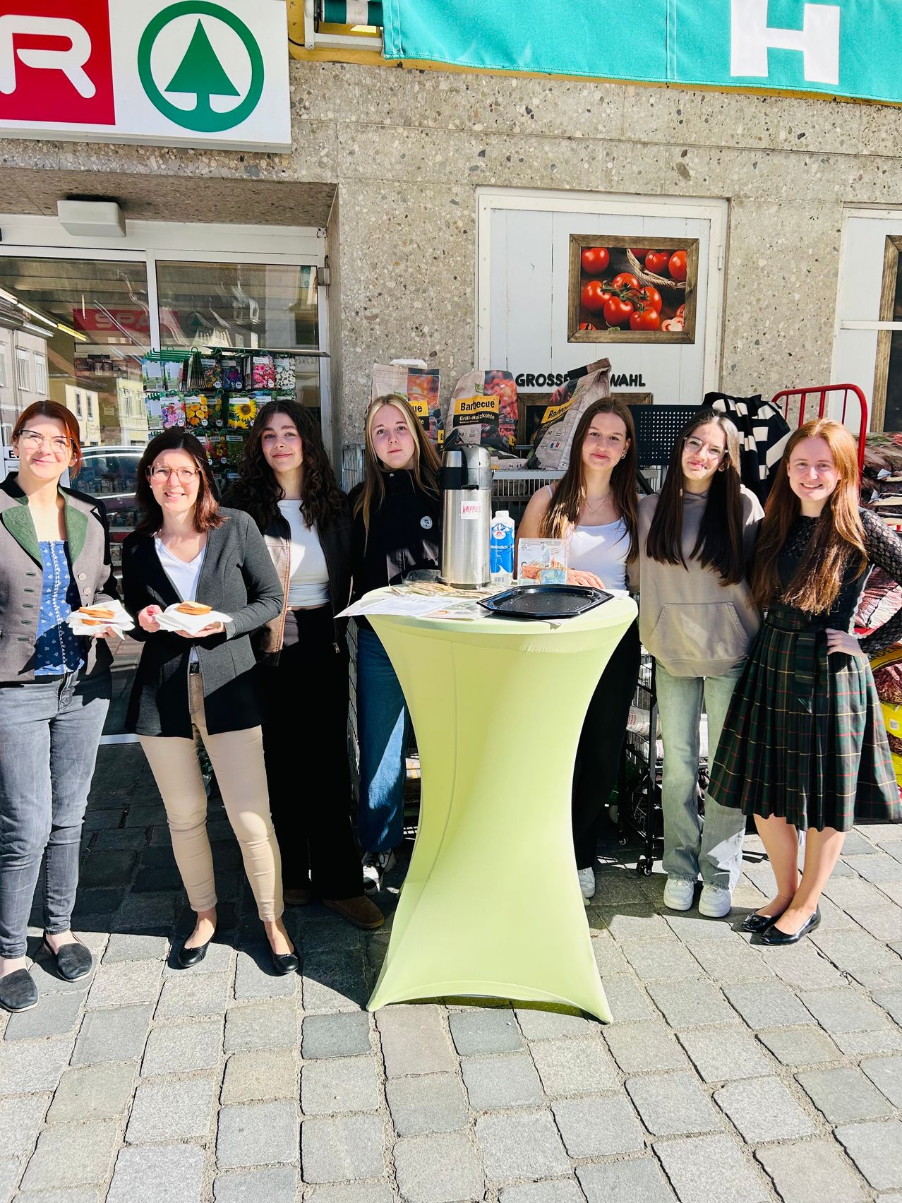 Eine Gruppe von Frauen steht draußen in der Nähe eines Tisches, lächelt und hält Essen. Ein Poster mit Tomaten ist hinter ihnen.