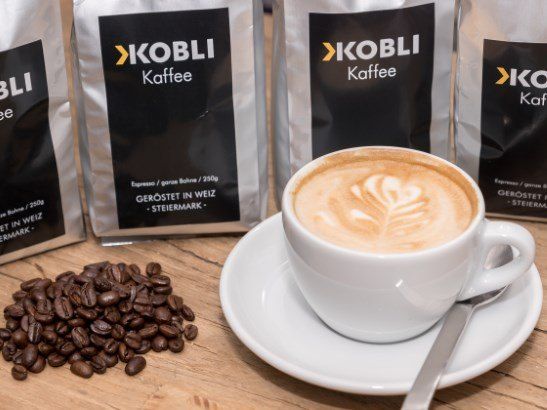 A cup of coffee with a leaf design on top sits on a saucer with a spoon. Behind it are bags of coffee labeled KOBLI.