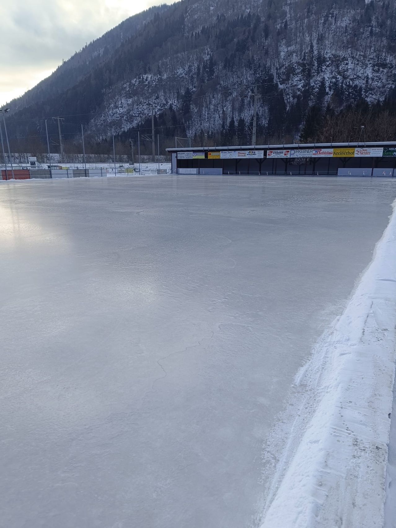 Eine große Eisbahn umgeben von einem verschneiten Berg, mit einem Gebäude im Hintergrund, das verschiedene Werbungen anzeigt.