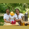 Eine Familie, bestehend aus einem lächelnden Mann, einer Frau und einem Kind, sitzt auf einem Holztisch mit Bienenstöcken im Hintergrund. Das Kind trägt rote Schuhe und hat einen smiley Gesichtsfilter.