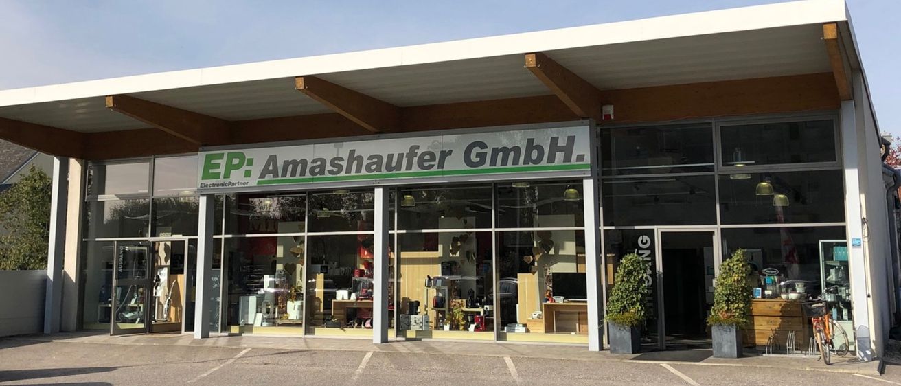 Die Fassade des EP: Amashauer GmbH-Geschäfts ist modern, mit Glaswänden, Holzbalken und Begrünung. Das Schild steht für EP: Amashauer GmbH, und verschiedene Artikel sind im Inneren ausgestellt.