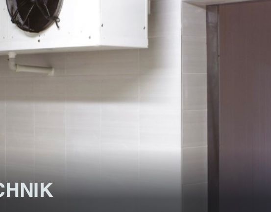 Eine gewerbliche Kühleinheit, die an der Wand über einer gefliesten Wand montiert ist, mit einer geschlossenen Tür auf der rechten Seite. Die Einheit hat einen schwarzen Lüfter und eine Rohrleitung an der Vorderseite. Das Bild hat einen Wasserzeichen, der 'ILTECHNIK' lautet.