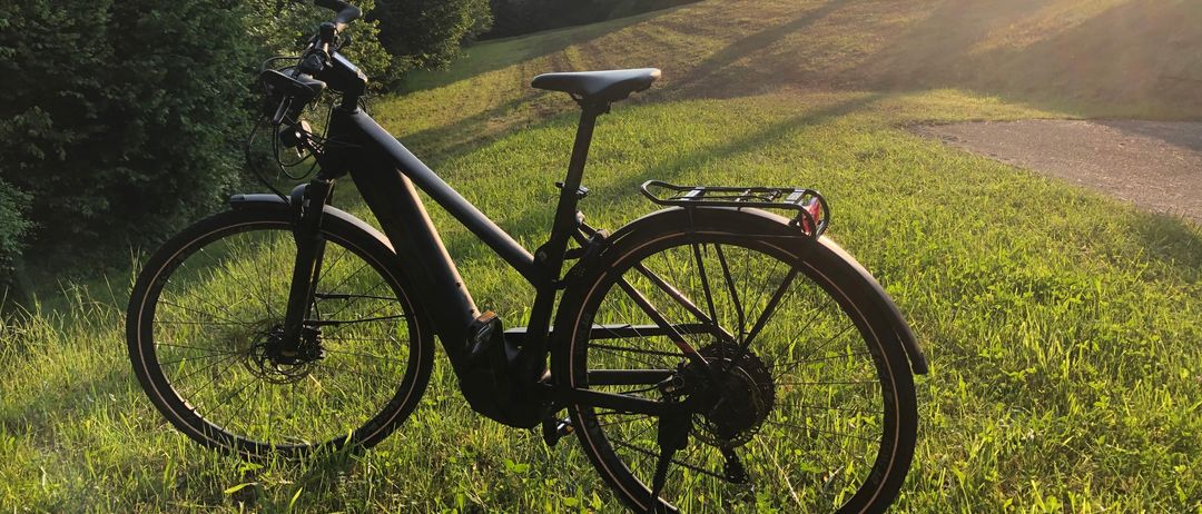 Ein schwarzes Fahrrad steht auf einem Grashügel, mit einem Gepäckträger an der Rückseite. Das Rad liegt mit eingefahrenem Stützfuß auf dem Boden. Im Hintergrund ist ein hügeliges Panorama mit Bäumen zu sehen.