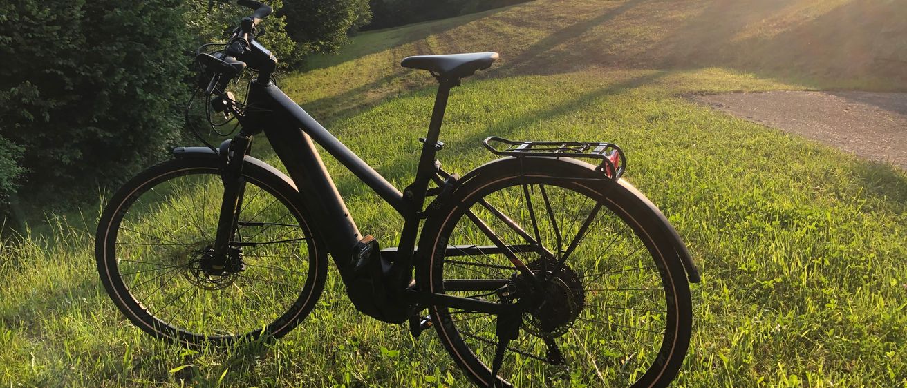 Ein schwarzes Fahrrad steht auf einem Grashügel, mit einem Gepäckträger an der Rückseite. Das Rad liegt mit eingefahrenem Stützfuß auf dem Boden. Im Hintergrund ist ein hügeliges Panorama mit Bäumen zu sehen.